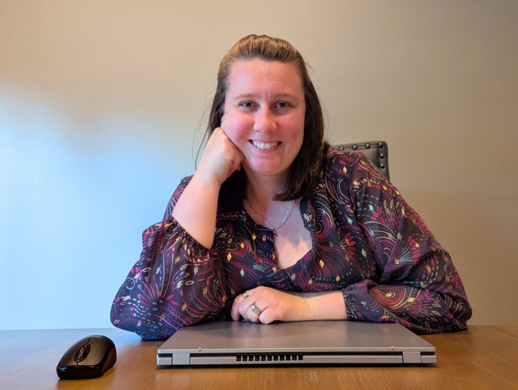 Alexandria Theordor smiles while seated at a desk with a closed laptop and a computer mouse in front of her. She wears a dark, patterned blouse with colorful abstract designs and rests her chin on her hand, giving a confident and approachable vibe against a plain beige background.