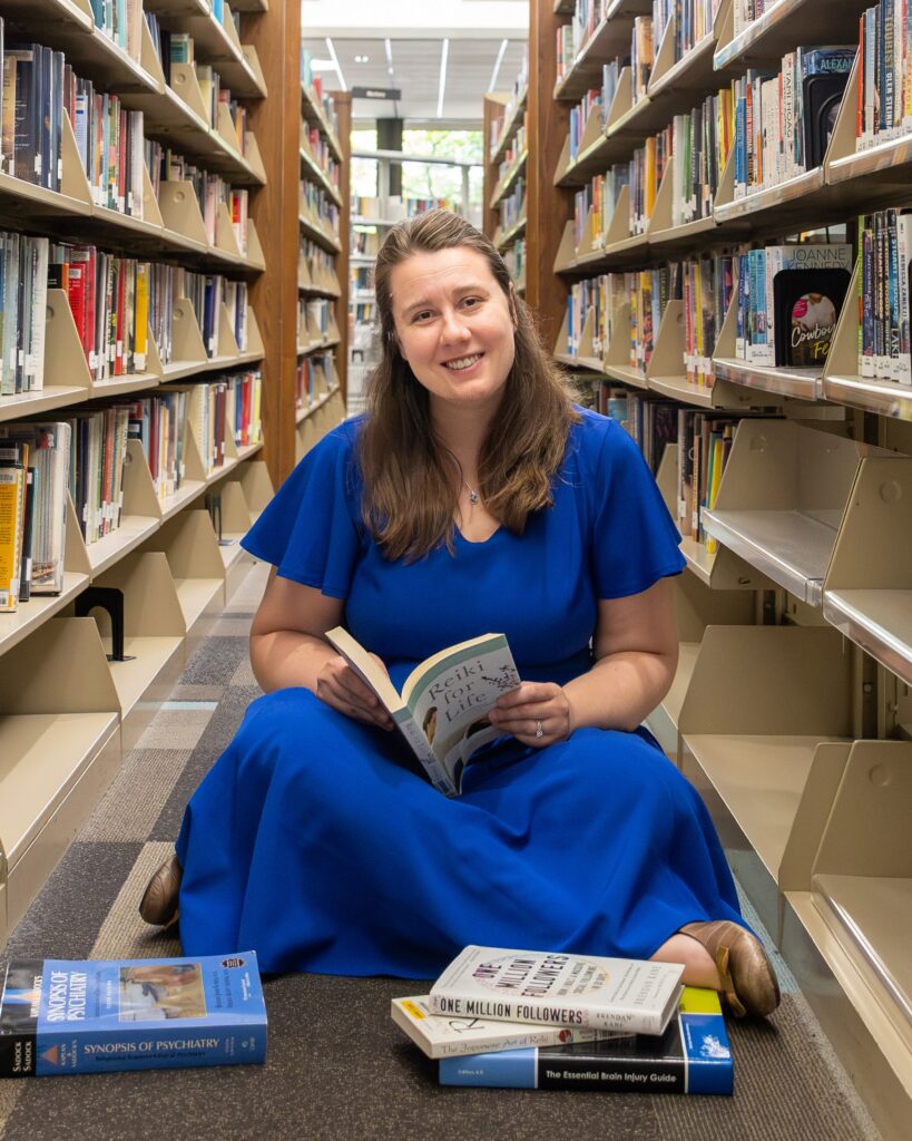 A woman in a bright blue dress sits on the floor between library shelves, smiling while reading a book. Several other books are stacked on the floor around her. | stresslessdigitalmarketingagency.com