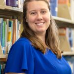 A woman with long brown hair, wearing a blue dress, smiles while standing in a library aisle with bookshelves in the background. | stresslessdigitalmarketingagency.com